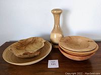 Photo showing a set of 7 wood bowls in various shapes and sizes along with a light wood vase on a wooden surface against a white background.