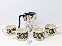 Photo showing clear flameproof glass carafe with black handle and gold wheat & thistle design surrounded by four beige speckled stoneware mugs with green leaf pattern.