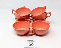 Top-down view of six salmon pink Melmac cups stacked in two groups, with the sugar and creamer pieces in front, all on a white surface.