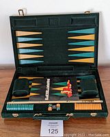 Open green velvet backgammon case showing playing surface with gold and teal points. Inside are two green velvet dice cups, stacks of green and orange checkers, five dice including two doubles, and a black instruction booklet labeled 'Backgammon'.