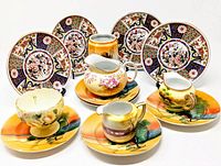 Various vintage fine china plates, mugs, creamers, and a small vase arranged in cluster showing detailed painted designs and colors.