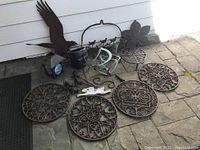 Overview of lot items arranged on stone patio including cast iron medallions, metal garden hooks, pottery head planter, metal eagle, and lantern