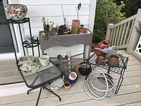 Photo showing the plastic resin wicker-look rectangular plant stand with multiple small pots, orchids, and a Southwest style pottery vase on a green metal plant stand; nearby metal tables hold watering cans and garden tools; garden hose coiled underneath.