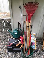 Photo showing assortment of garden tools including rakes, hand tools, watering can, and unopened fertilizer bags.