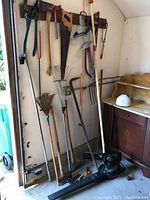 Photo of various garden and shop tools against a wall including a hedge trimmer, saw, rakes, and blower.
