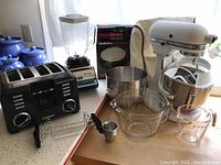 Various kitchen appliances including KitchenAid mixer, waffle maker, blender, toaster, and glass measuring cups on a wooden table