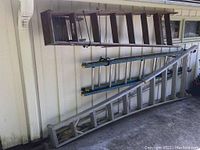 Three ladders hanging on house wall: wood ladder top, green Werner extension ladder middle, silver metal ladder bottom