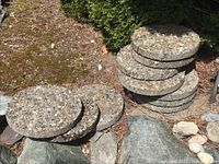 Stack and spread of round aggregate concrete stepping stones about 14 inches diameter and 2 inches thick with embedded stones on surface.