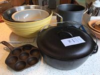 Overview of all items in the lot showing the Lodge cast iron pot with lid and handle, yellow pottery bowl, stainless steel bowl, Texas Ware plastic bowl, black metal stock pot, and vintage Jotul cast iron muffin tin