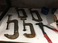 Photo of five large C-clamps and one rivet tightener tool arranged on a white surface.