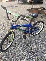 Kids Magna bicycle with blue frame and green fork, black padded seat with skull design, black handlebars with green grips, white tires on gravel surface.