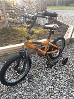 Front side view of the orange kids Mongoose bicycle with training wheels, showing rust and wear on the frame and seat.