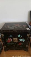 Front view of the black Asian side table showing hand-painted floral and bird design on doors and brass hardware.