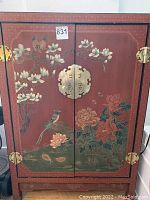 Front view of red lacquer cabinet, two doors closed showing painted trees, birds and peonies, brass latch and hinges