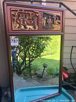 Front view of the wall mirror showing the rectangular mirror and the detailed three-dimensional carved panel at top with Asian style figures in gold paint.