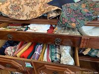 Drawer and surfaces filled with assorted table linens, including folded fabrics, lace napkins, and tapestry-style textiles