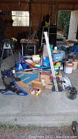 Garage floor view showing jack stands, floor jack, bike rack, roof rack bars, cleaning supplies, and various accessories on cardboard and floor.