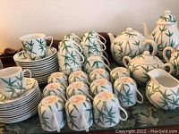Full view of 12 teacups and saucers, 12 demitasse cups and saucers, 2 cream and sugar sets, coffee pot and teapot decorated with green bamboo motif on white porcelain.