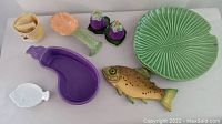 Full view of ceramic items on white surface including vegetable and plant-themed plates, salt and pepper shakers, fish plate, and egg separator.