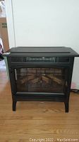 Front view of black Duraflame electric fireplace insert with glass front and faux logs visible inside.
