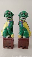 Front view of both ceramic Foo Dogs seated on brown rectangular bases, showing face details, painted colors and spheres under paws.