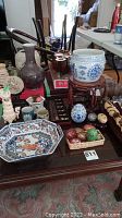 Overview of all items in lot on a table featuring porcelain ginger jar on wooden stand, Kashmiri eggs on wooden tray, wooden carved masks, chopsticks, and assorted vases and figurines.