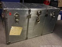 Front and top perspective showing metal plating, riveted construction, corner reinforcements, and three central locking mechanisms. Yellowed paper label affixed to side.