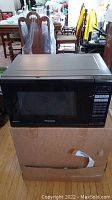 Black Panasonic microwave placed on a cardboard box in a room with wooden flooring and furniture in the background, showing the front and top of the microwave.