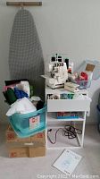 Singer serger atop white two-tier table, ironing board behind, bins of fabric and notions below