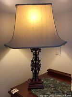 Full view of the table lamp illuminated showing the fabric shade and carved wooden base with Asian motifs.