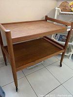 Front angle view of teak trolley showing two shelves and casters on legs.