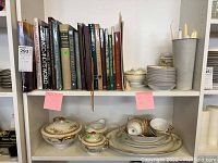 Shelf shows two rows: top row with coffee table books, bottom row with china dinnerware pieces including serving platters, bowls, covered sugar bowl.