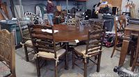 Round wooden dining table surrounded by six ladderback chairs with cane seats and cushions. The space includes other stored furniture in background.