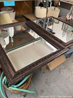 Two large framed mirrors displayed on a surface with reflections of lamps and shelving. Both frames are dark wood with decorative carving around edges.