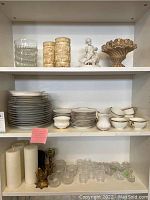 Photo showing top three shelves with glassware including bowls, vases, porcelain china with gold rim, white pillar candles, brass candle holders, and glass candle holders.