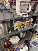 Photo shows multiple kitchen appliances and boxed items on metal shelves including coffee maker, coffee urns, ice cream maker, tea kettle, and other utensils.