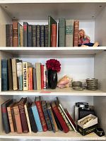 Three shelves with assorted vintage and classic hardcover books, a doll, vintage photo equipment, vases, coasters, decorative items