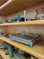 Three wooden middle shelves containing assorted silverplate serving ware and decorative kitchen items including trays, covered dishes, candlestick, and pots.