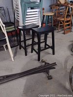 View of two black wood square seat counter height stools placed on concrete floor surrounded by other stacked chairs and miscellaneous items.