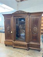 Front view of vintage wooden wardrobe showing the three main doors, central mirror, and carved floral wood details on each side door and top crown.
