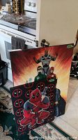Two acrylic paintings, one featuring several Marvel characters including Deadpool, Green Lantern, Spider-Man, and Cyborg, predominantly red and black with comic style.
