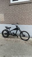 Left side view of all black low rider bicycle showing full frame, tires, pedals and saddle against brick wall