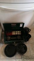 Full view of Coleman green two-burner stove with black enamel cookware arranged around it on floor
