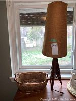 Full view of the wooden tripod lamp with tall burlap shade and the wicker basket beside it on a wooden surface near a window.