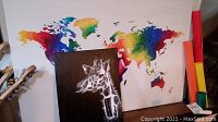 Wide shot showing both paintings together: the large colorful world map backdrop and the smaller black canvas with white giraffe portrait in front.