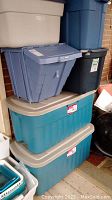 Multiple large plastic storage bins stacked, showing colors teal, beige, blue, black and gray with lids or without.