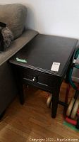 Dark wooden side table next to a gray couch with a drawer front and round knob, visible minor scratches on surface.