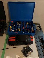 Overview of blue plastic carrying case filled with multiple miniature Hot Wheels vehicles and small toy construction vehicles. Black remote control car with red stripes placed in front alongside controller.