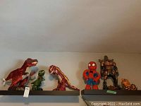 Front view showing the six toy figures lined up on shelves against a wall. The large Tyrannosaurus rex plush is on the left side, next to two smaller dinosaur figures, then Spider-Man, Grimlock Transformer, and the cat figure on the right.