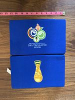 Top and bottom view of one blue seat cushion featuring 2006 FIFA World Cup logo and trophy graphic, alongside measuring tape showing size.